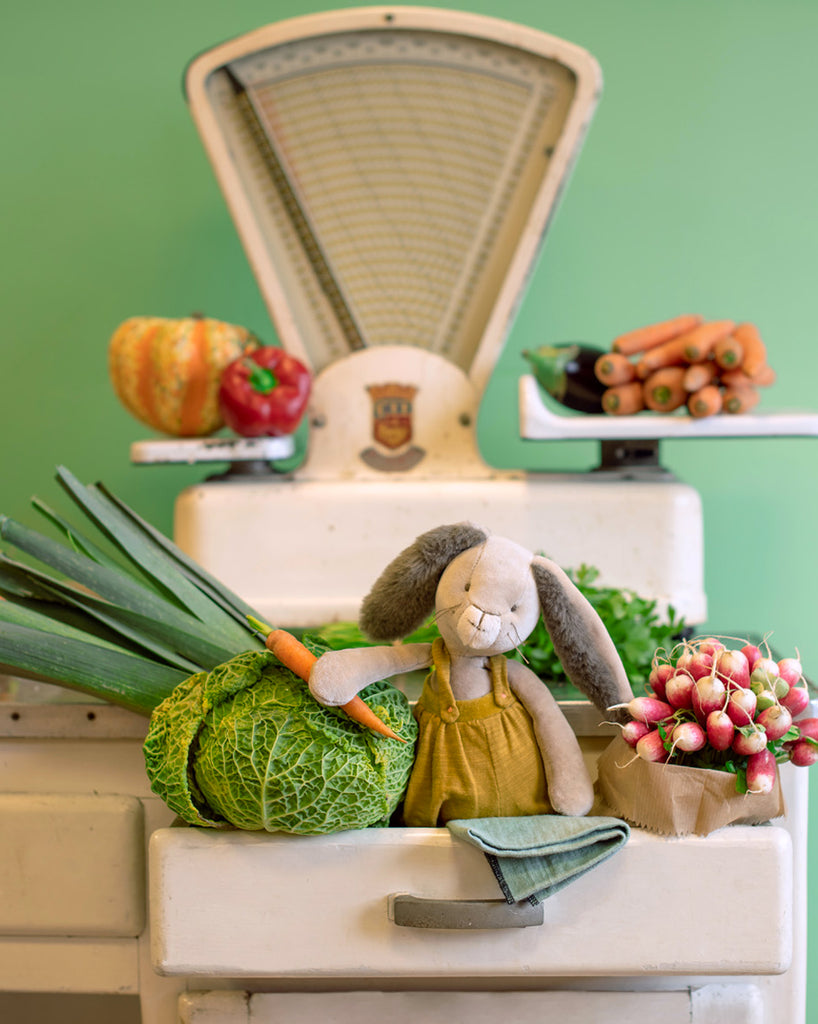 Ochre Petit Rabbit from Moulin Roty’s Trois Petits Lapins collection—seated in a sink holding a carrot, surrounded by cabbages, radishes & vintage scales. Available at Michelle Kindness Design.